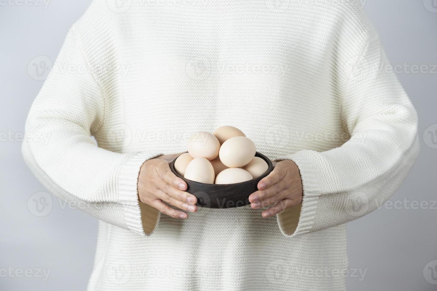 Mains féminines tenant un bol en bois plein d'oeufs de poule crus contre un mur gris photo