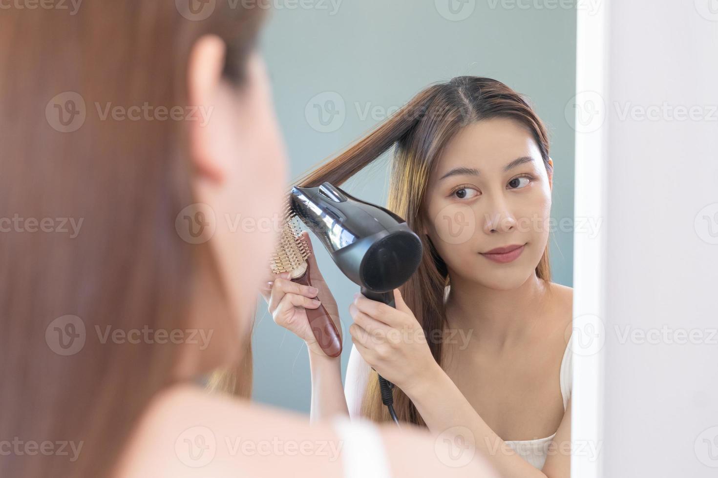 cheveux séchoir, content, asiatique Jeune femme, fille à la recherche dans miroir réfléchir en utilisant sèche-cheveux à sec, soufflant longue blond tout droit après douche à maison. coiffure, cheveux traitement et beauté santé se soucier. photo