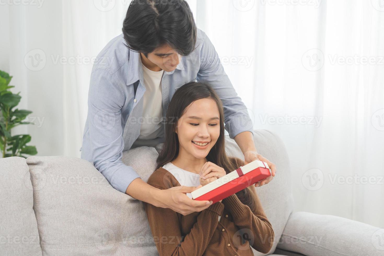 célébrer la Saint-Valentin journée anniversaire, relation deux asiatique Jeune couple aimer, copain donne une cadeau à petite amie par cacher boîte à le derrière, femme avoir présent tandis que séance sur canapé à maison. photo