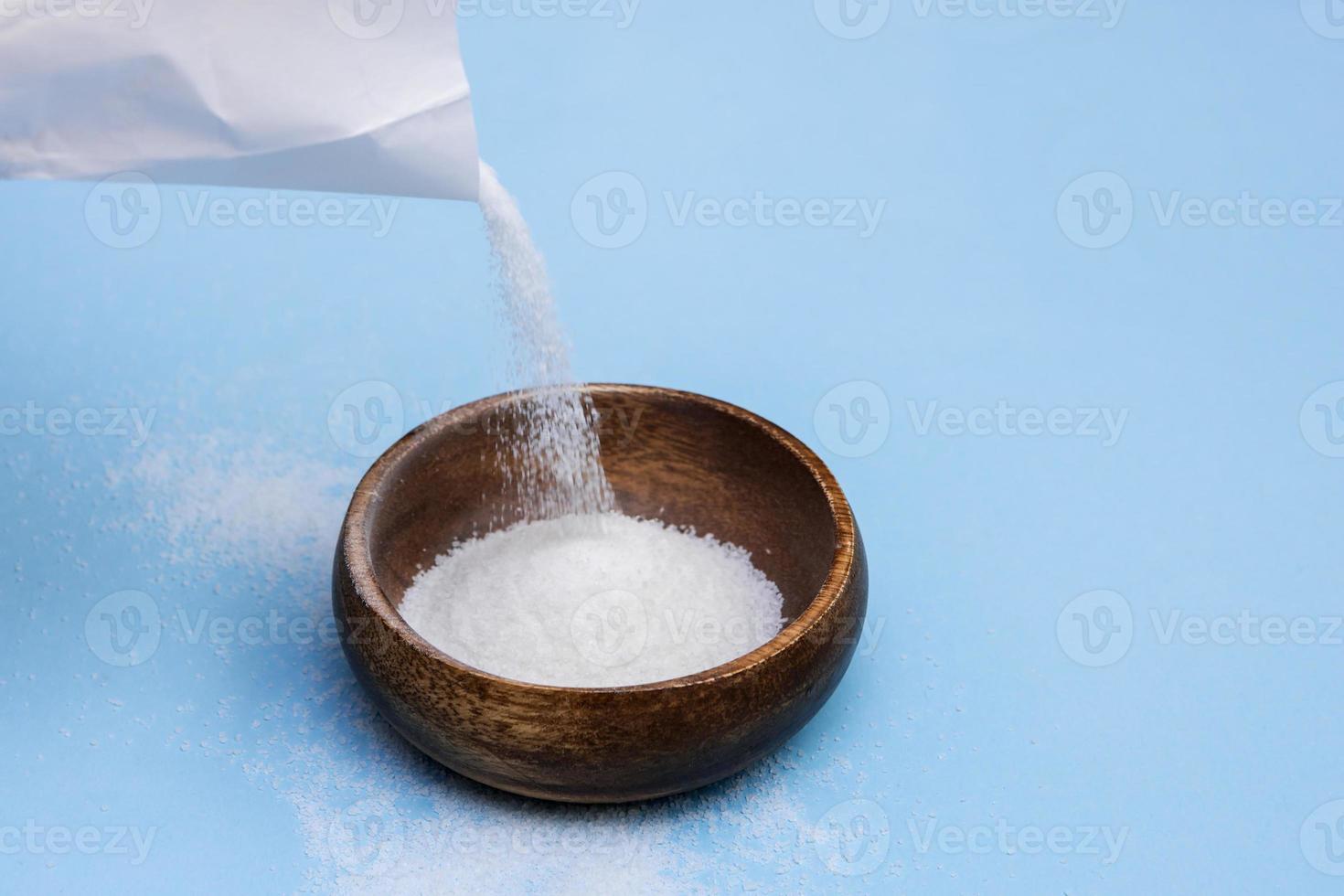 sucre est versé de une blanc papier sac dans une en bois bol sur une bleu Contexte photo