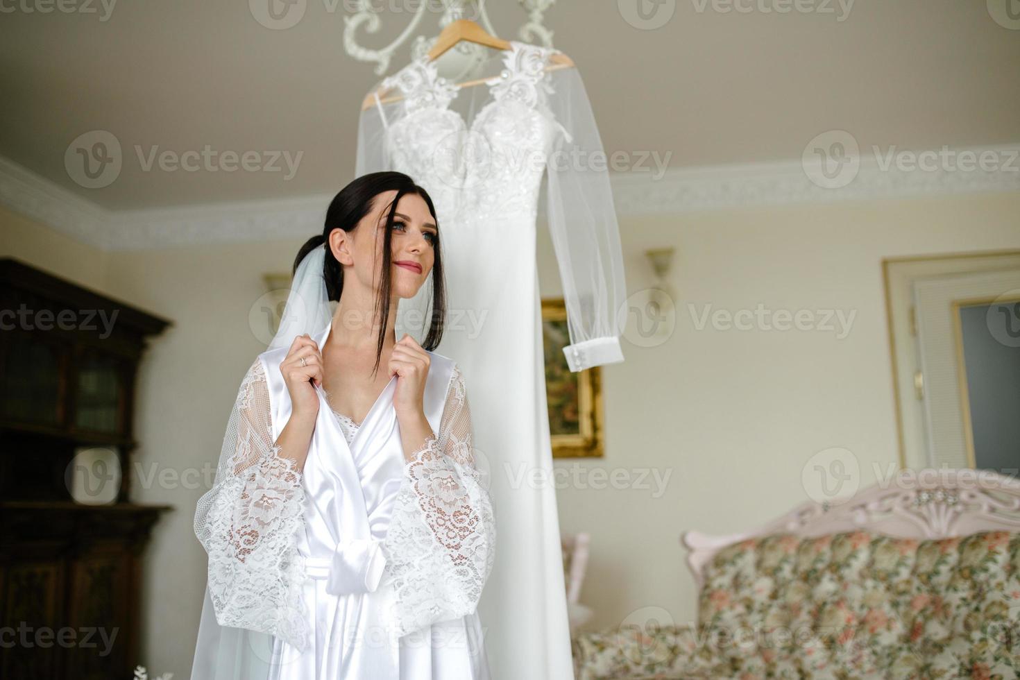 le Matin de le la mariée. portrait de une brunette la mariée dans une blanc soie manteau derrière une robe photo