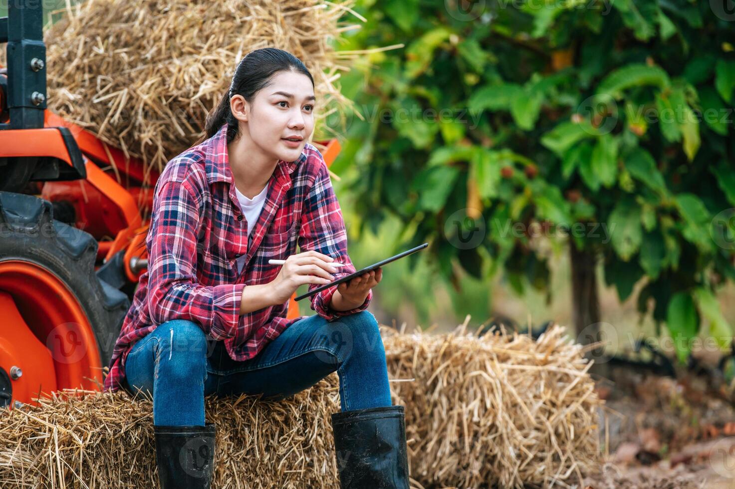 jeune agricultrice asiatique assise sur une balle de foin avec un gros tracteur dans les terres agricoles. technologies modernes dans la gestion de l'agriculture et le concept de l'agro-industrie. photo