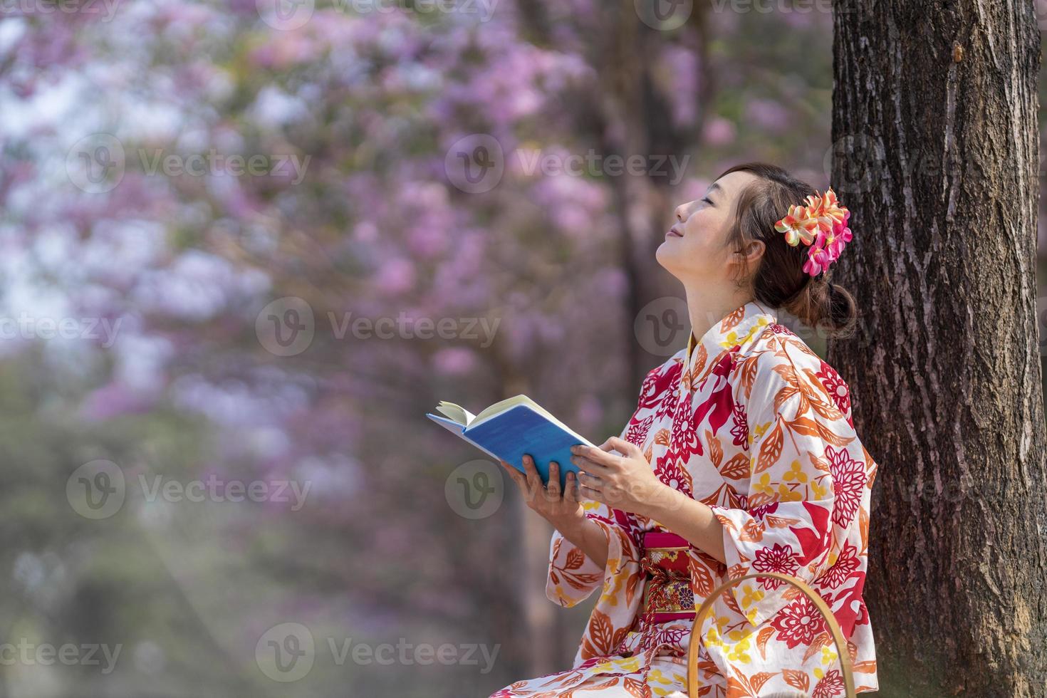 Japonais femme dans traditionnel kimono robe séance en dessous de Cerise fleur arbre tandis que en train de lire une livre pendant printemps Sakura Festival concept photo