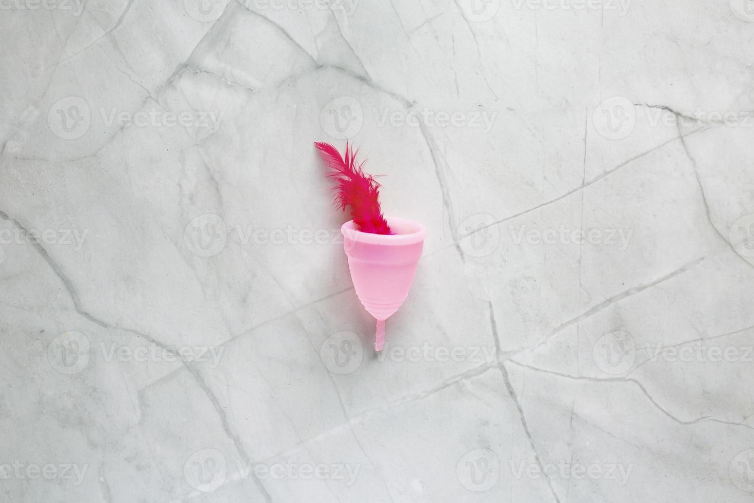 menstruel tasse et rouge oiseau plume sur marbre Contexte. alternative féminin hygiène produit pendant le période. femmes santé concept photo