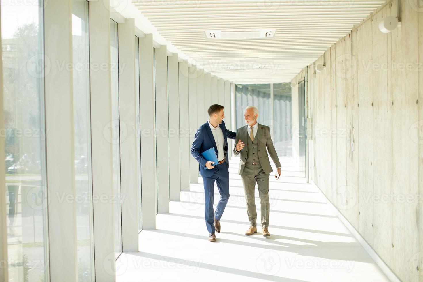 Jeune et une Sénior homme d'affaire marcher vers le bas un Bureau couloir, Profond dans conversation photo