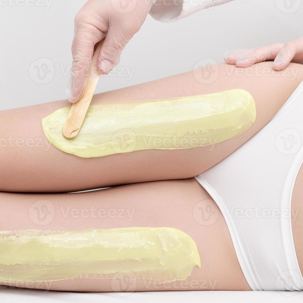 cosmétologue dans gants s'applique chaud la cire sur svelte femme jambes en utilisant spatule. épilation dans beauté salon photo