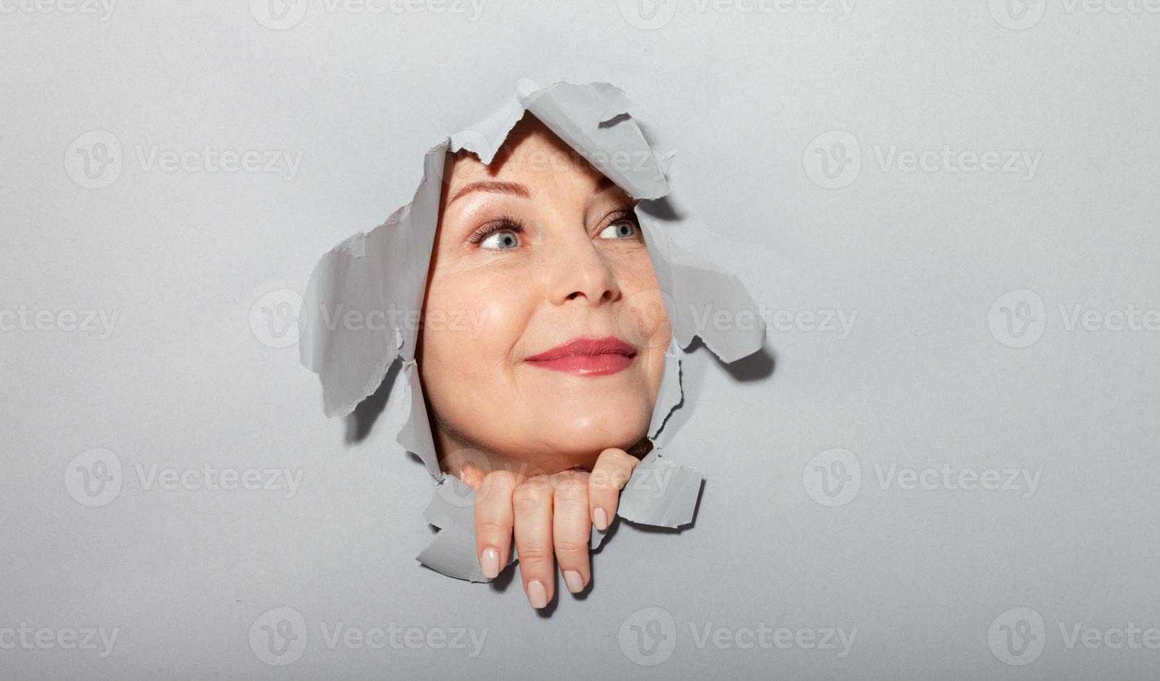 femme surprise regardant de manière ludique dans un trou de papier déchiré, a excité une expression joyeuse, regarde à travers une percée de fond gris. waouh super nouvelle photo