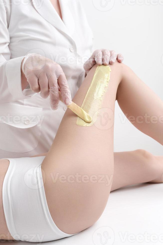 méconnaissable esthéticien dans blanc manteau appliquant vert chaud la cire sur femme jambe en utilisant spatule photo