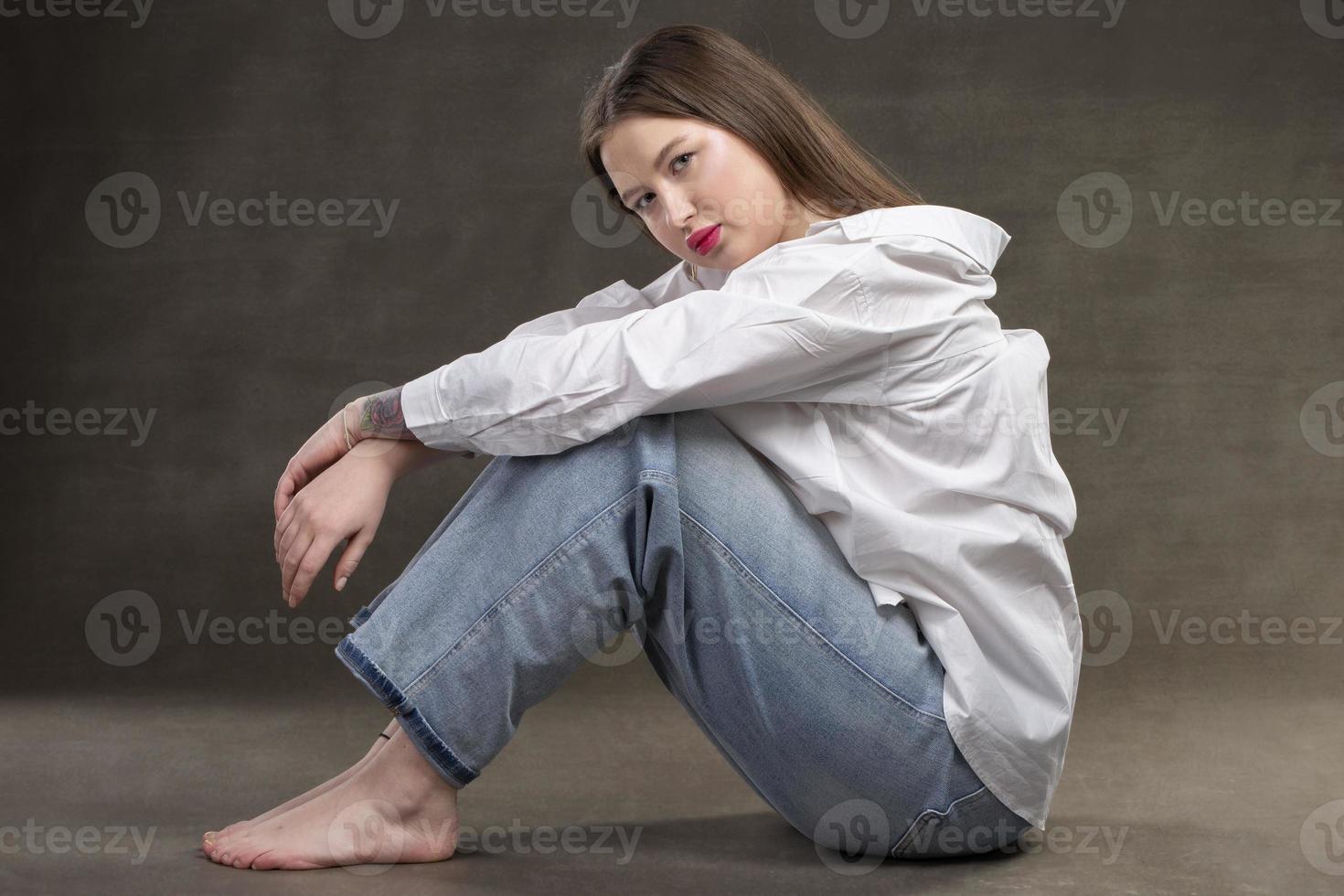 une magnifique fille dans une blanc chemise et jeans est assis sur une gris Contexte. photo