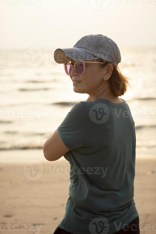 portrait de asiatique femme portant jeans casquette permanent à mer plage contre magnifique le coucher du soleil lumière photo