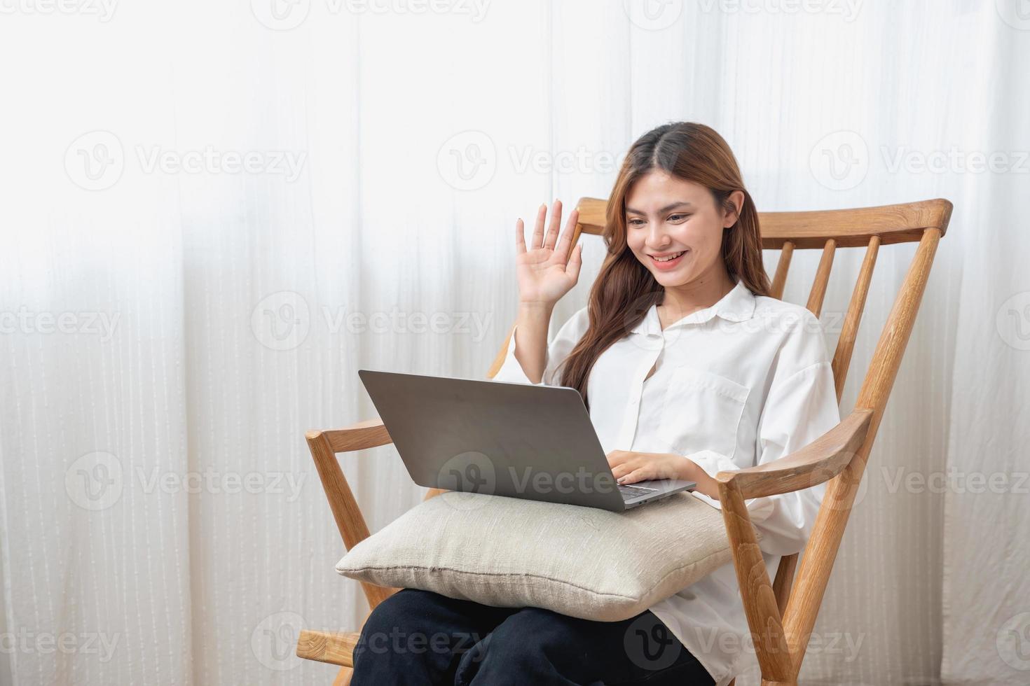 portrait de une magnifique Jeune femme portant une blanc chemise en portant une portable à type sa posséder récit et séance sur une en bois chaise à l'intérieur le loger, le concept de du repos et relaxation photo