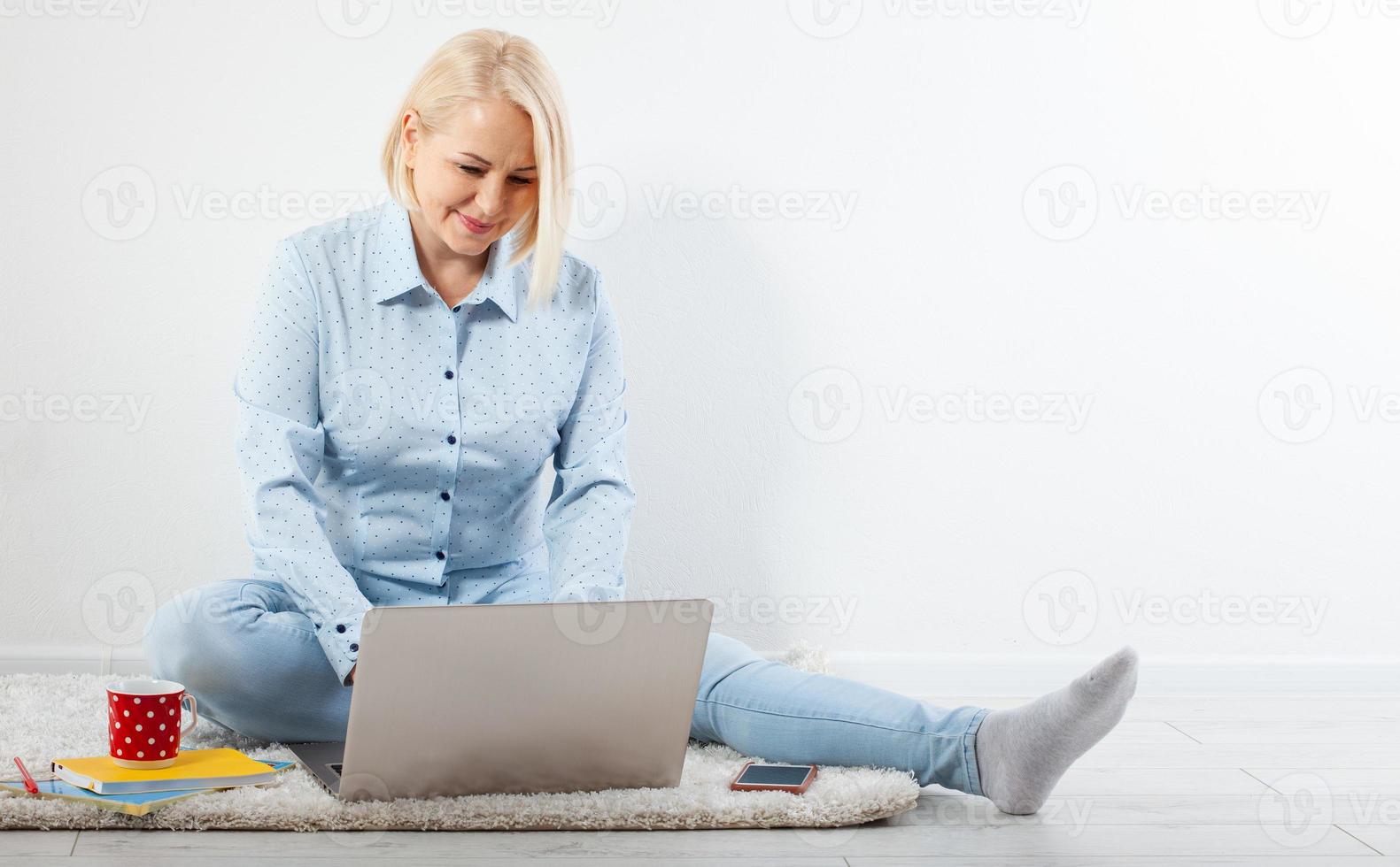 content milieu vieilli femme séance détendu sur le sol en utilisant portable pour divertissement. le concept de loisir et travail avec une tasse de café à maison. photo