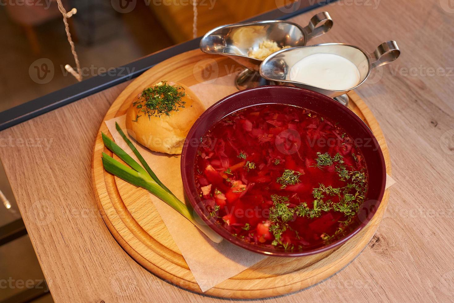 Une photo d'un bortsch ukrainien avec une pampushka, l'oignon vert et la crème sure sur un plateau en bois sur la table