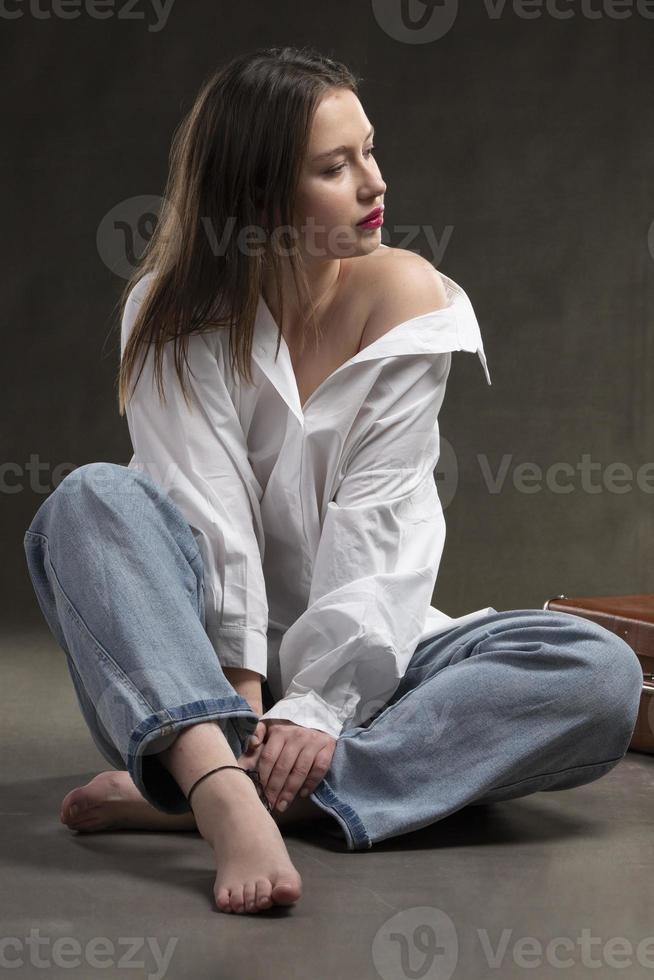 une magnifique fille dans une blanc chemise et jeans est assis sur une gris Contexte. photo