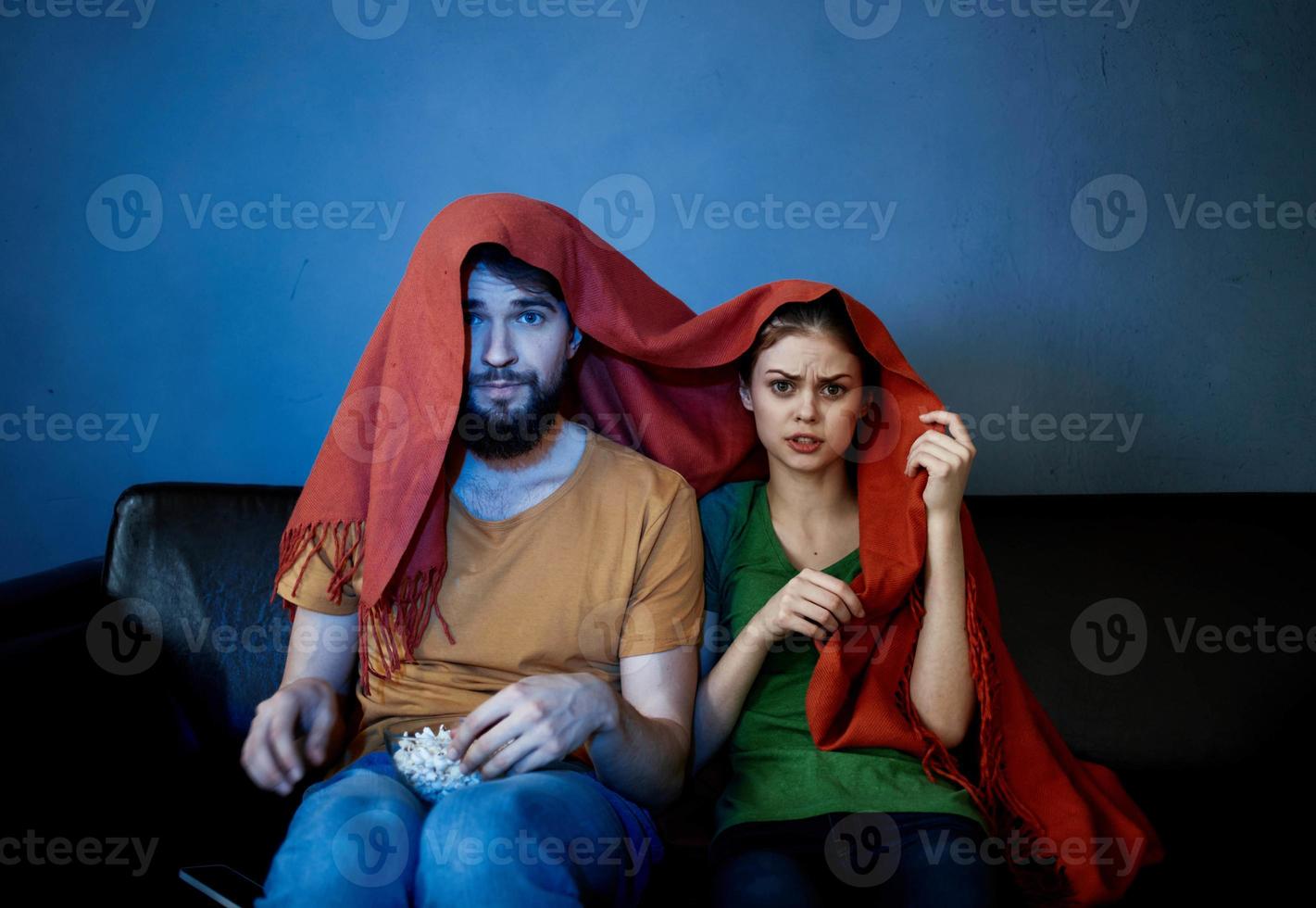une femme et une homme en dessous de une rouge couverture sur le canapé en train de regarder la télé dans le soir photo