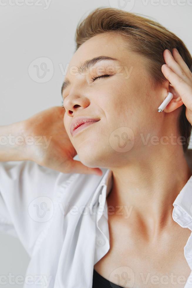 Jeune femme adolescent écoute à la musique avec infertile écouteurs et dansant maison, sourire avec les dents avec une court la Coupe de cheveux dans une blanc chemise sur une blanc Contexte. fille Naturel pose avec non filtres photo