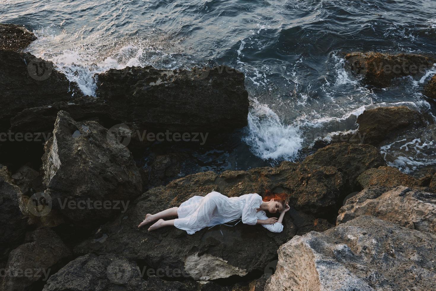 magnifique femme dans longue blanc robe humide cheveux mensonge sur une rocheux falaise vacances concept photo