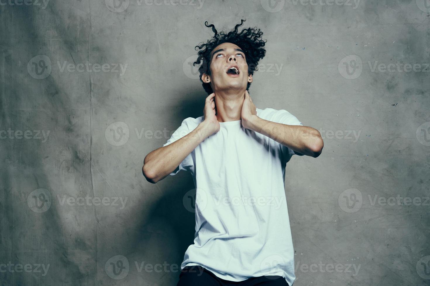 de bonne humeur gars avec frisé cheveux gestes avec le sien mains près le sien visage sur une gris en tissu Contexte dans une blanc T-shirt photo