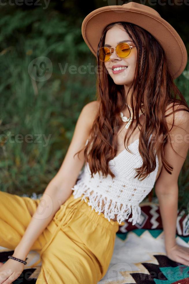 une Jeune hippie femme dans éco-vêtements a amusement en riant et servile séance sur une plaid à l'extérieur dans le tomber avec une chapeau et Jaune des lunettes de soleil. Voyage mode de vie photo