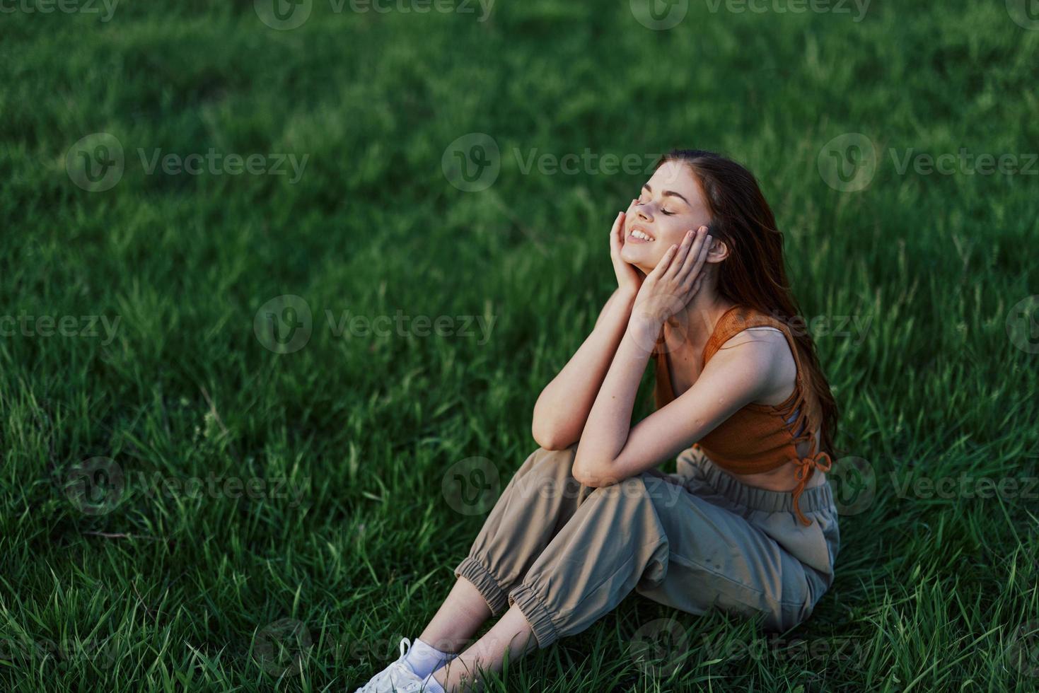 une magnifique fille relaxant dans la nature séance dans un pantalon et Haut sur le vert herbe dans le des rayons de le réglage été Soleil. le concept de santé et se soucier pour le corps et le environnement photo