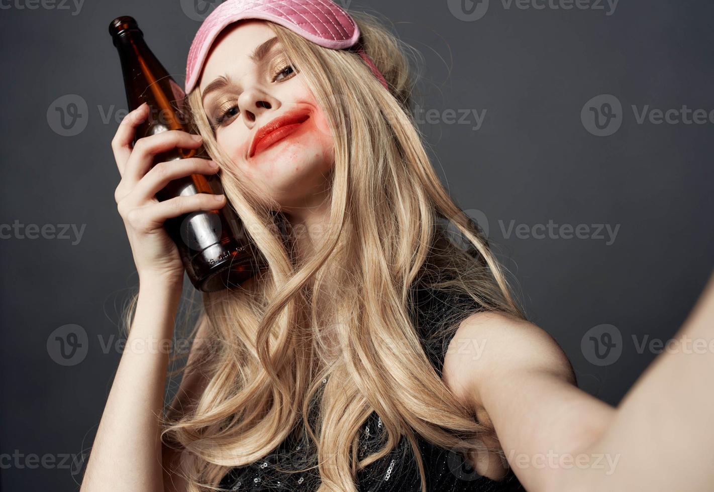 ivre femme avec une bouteille de Bière sur une gris Contexte gestes avec sa mains et brillant maquillage photo