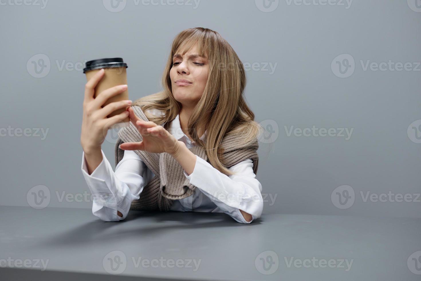 malheureux irrité Jeune blond étudiant Dame pigiste dans chaud chandail Regardez à à emporter café séance à lieu de travail dans gris moderne Accueil bureau. café Pause amoureux concept. copie espace. cool offre photo