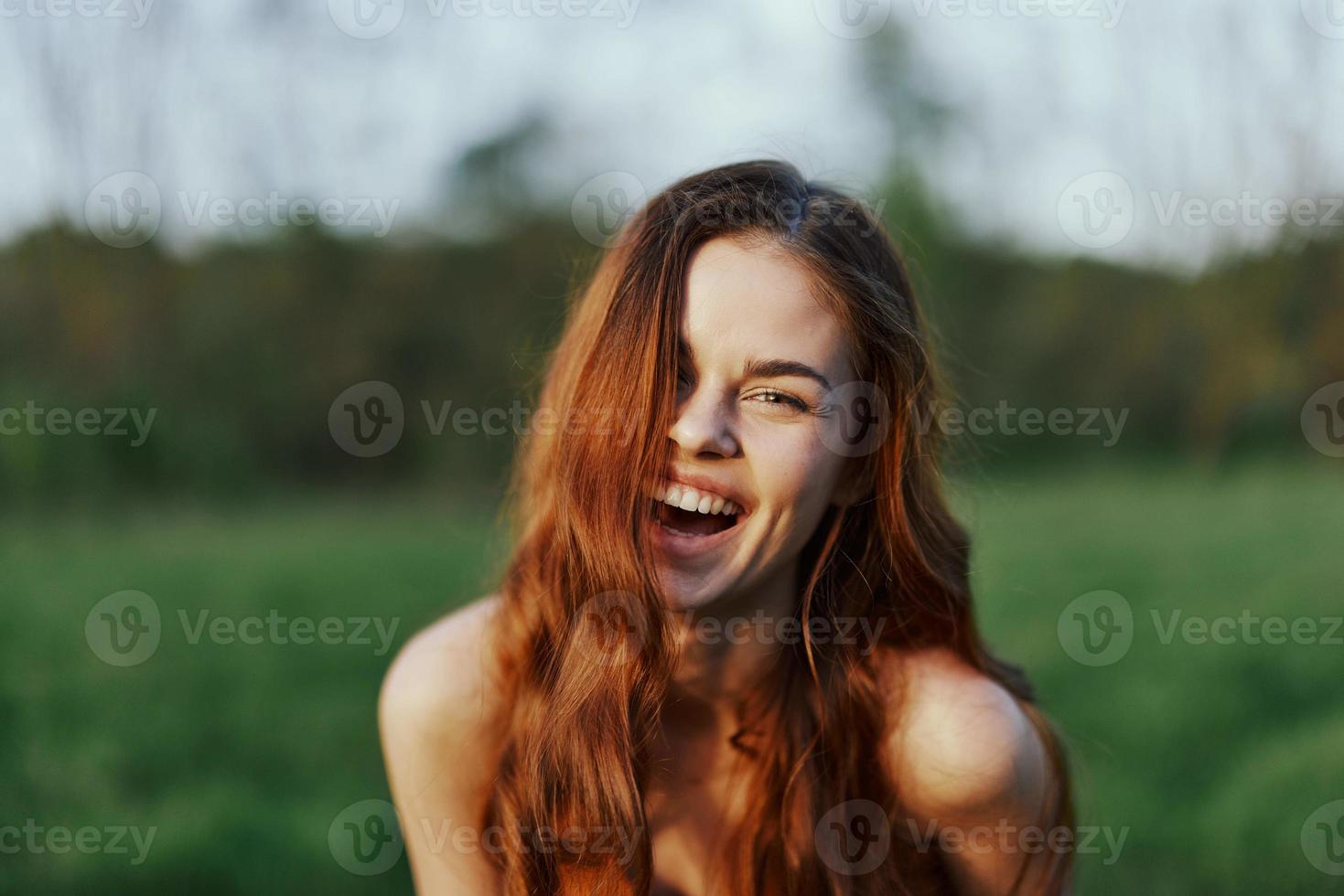 une Jeune femme sourit et regards dans le caméra avec sa long, rouge, ondulé, brillant cheveux dans une parc avec vert herbe dans le été le coucher du soleil. le concept de en bonne santé vie, beauté et cheveux se soucier. photo