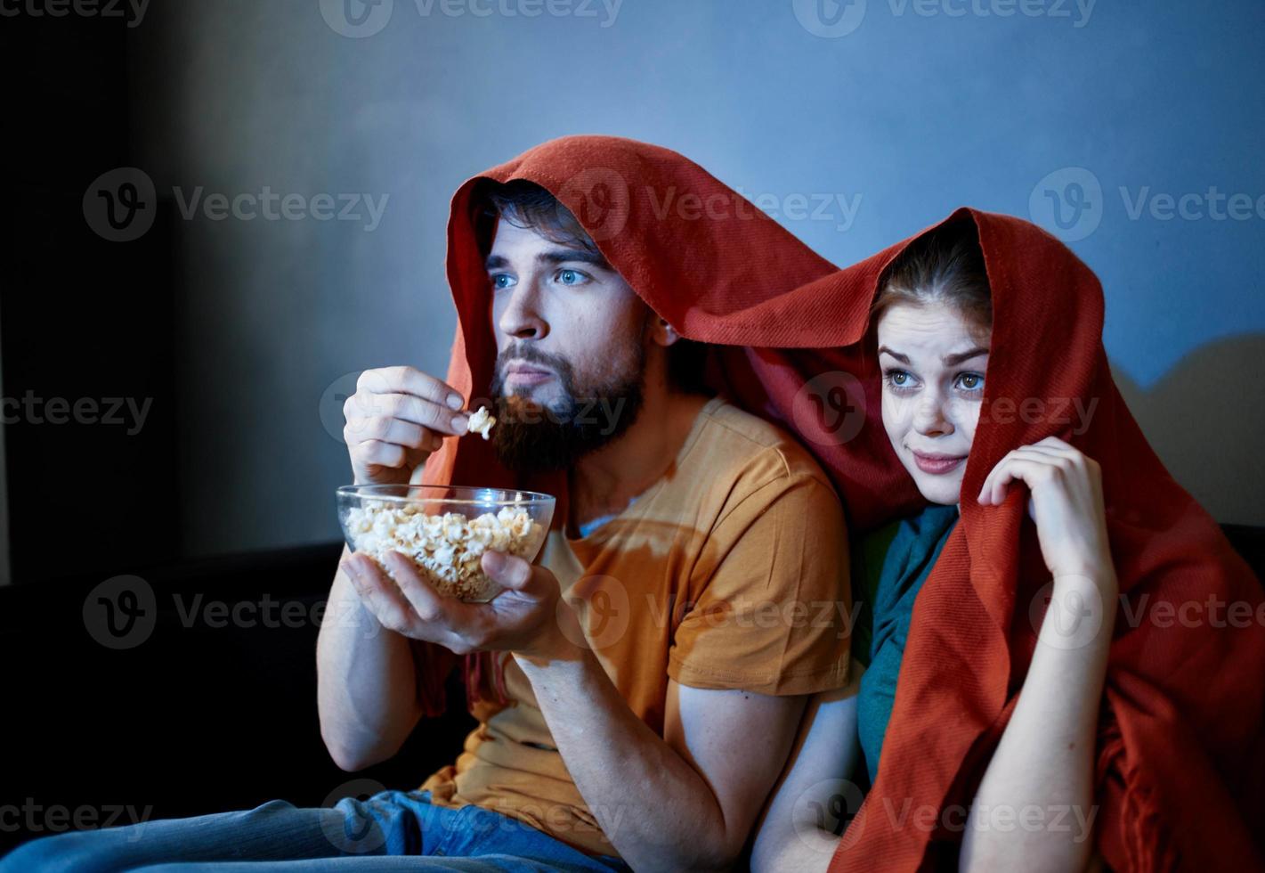 une homme avec une assiette de pop corn et un émotif femme en dessous de une rouge couverture sur le canapé photo