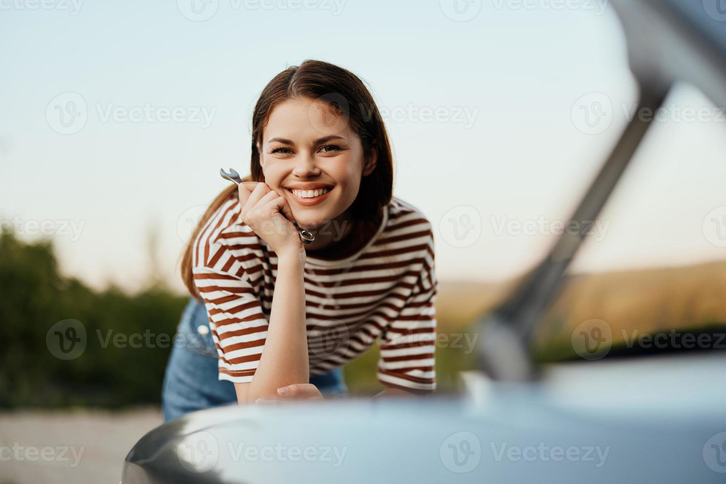 une femme avec une clé avec une sourire Heureusement regards en dessous de le ouvert capuche de sa voiture et réparations il de une bord de la route panne sur une route voyage seul photo