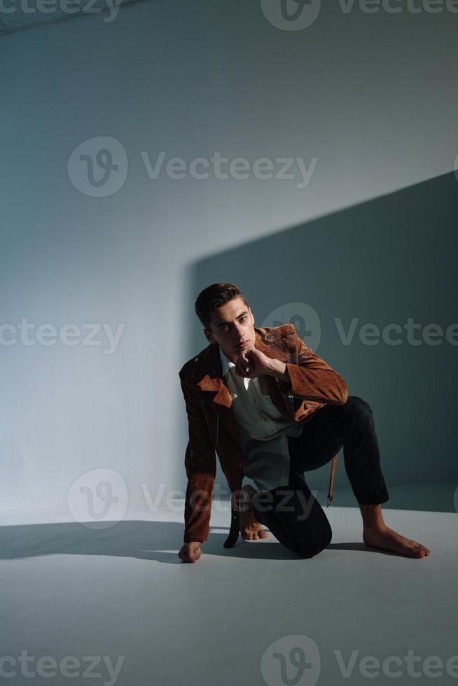 une gars dans une marron manteau et jeans est assis sur le sol dans une gris chambre. photo