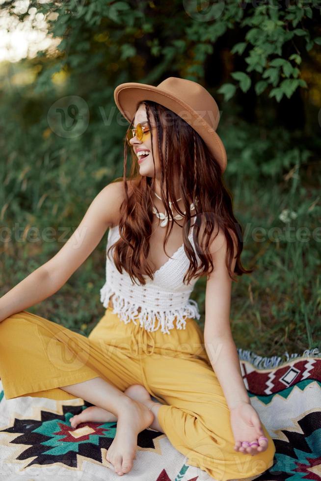 une Jeune hippie femme dans éco-vêtements a amusement en riant et servile séance sur une plaid à l'extérieur dans le tomber avec une chapeau et Jaune des lunettes de soleil. Voyage mode de vie photo