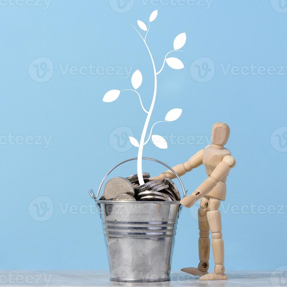 une en bois mannequin et une miniature seau avec métal pièces de monnaie et une peint arbre avec feuilles sur une bleu Contexte. le revenu et profit croissance concept photo