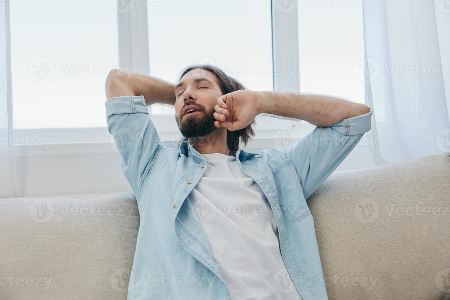 une homme est assis sur le canapé bâillement et élongation après une sieste, manquer de de sommeil et fatigue de travail et non conforme du quotidien routine. photo