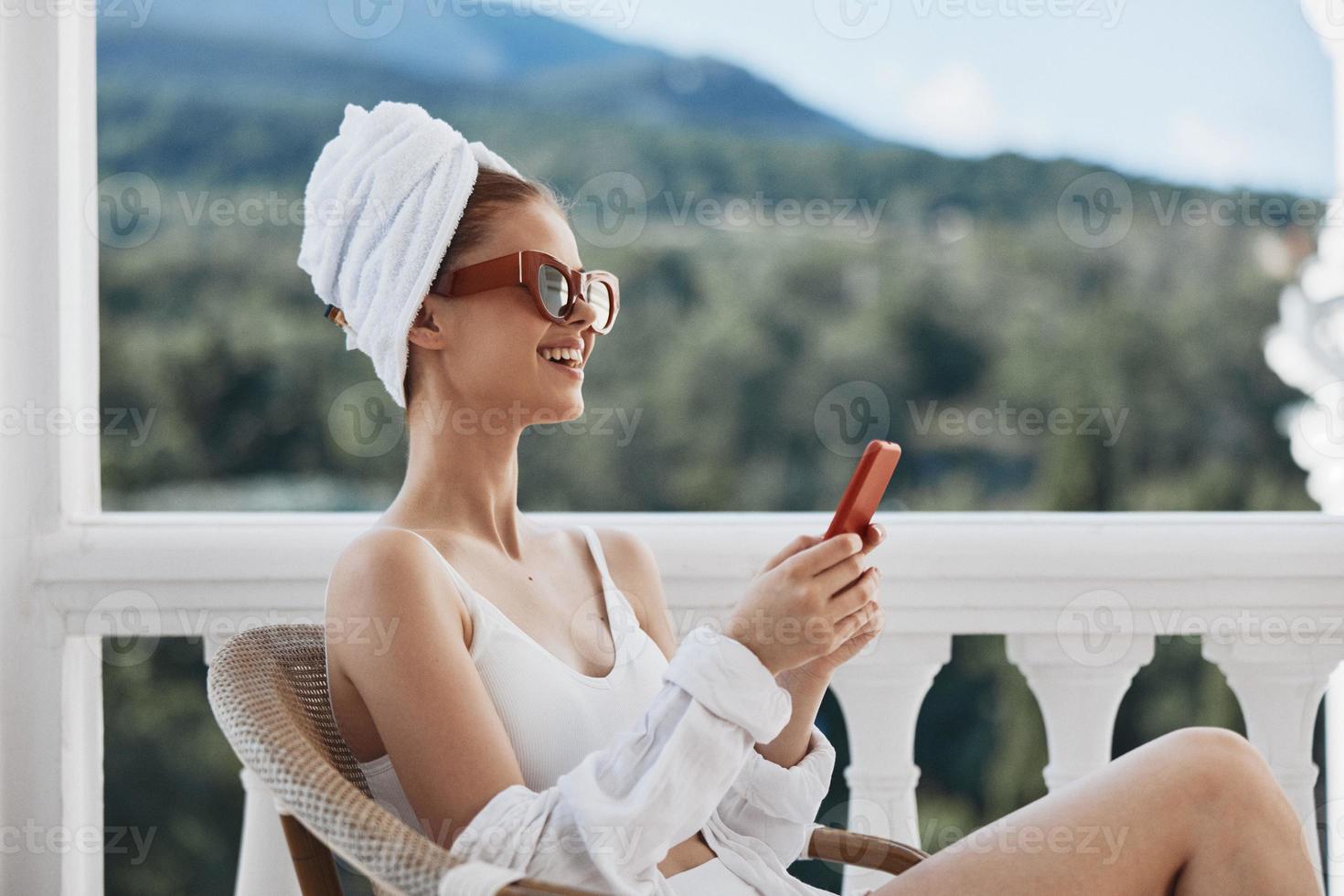 attrayant Jeune femme dans des lunettes de soleil est assis sur le balcon et les usages le téléphone paresseux Matin photo