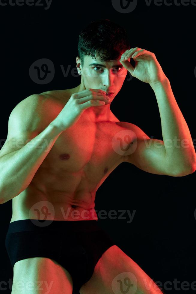 sexy Masculin bodybuilder gestes avec le sien mains sur une noir Contexte et culotte photo