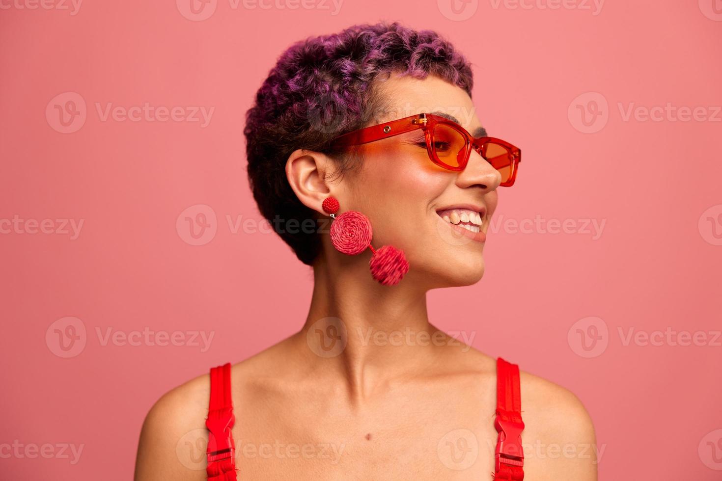 mode portrait de une femme avec une court la Coupe de cheveux dans coloré des lunettes de soleil avec inhabituel accessoires avec des boucles d'oreilles souriant sur une rose brillant Contexte photo