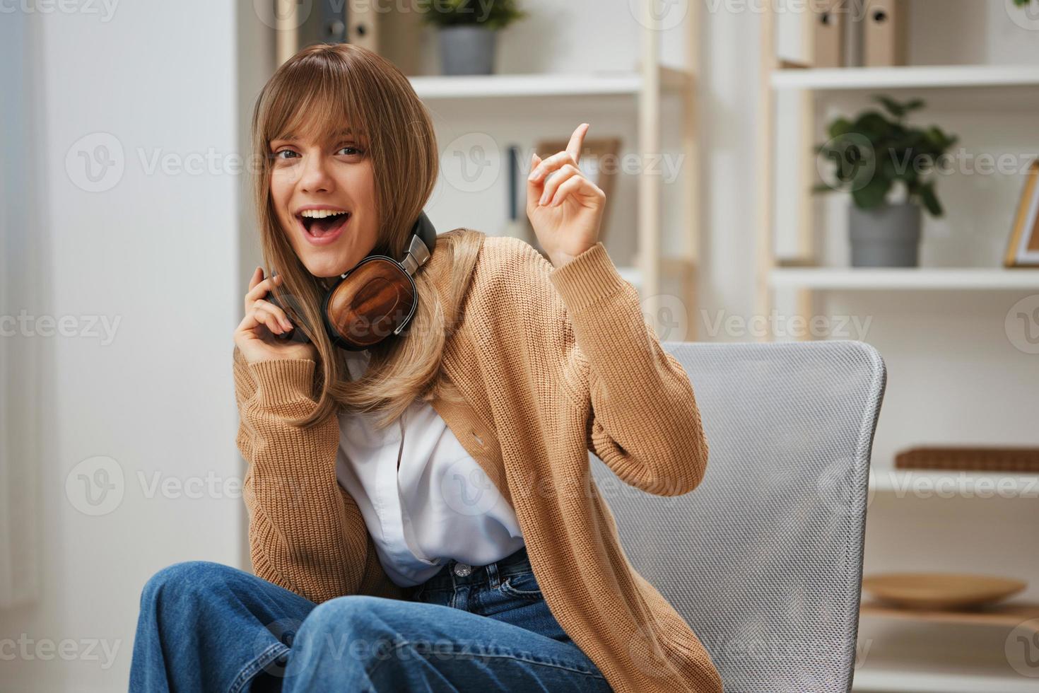 joyeux excité Jeune blond étudiant Dame dans chaud chandail dans écouteurs point doigt en haut à copie espace séance dans fauteuil à moderne Accueil intérieur. la musique temps relaxant cool playlist concept. cool offre photo