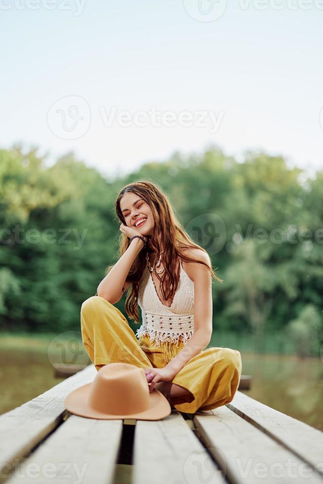 une Jeune femme souriant dans un image de une hippie et éco-vêtements séance en plein air sur une pont par une Lac portant une chapeau et Jaune un pantalon dans le été le coucher du soleil photo