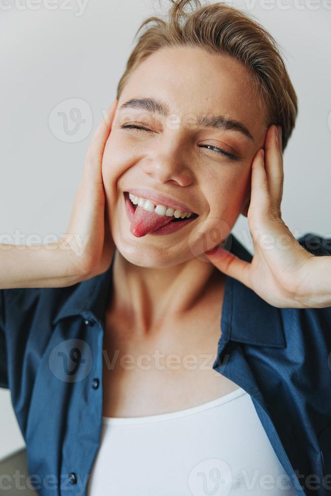 une Jeune femme séance dans une chaise à Accueil souriant avec les dents avec une court la Coupe de cheveux dans jeans et une denim chemise sur une blanc Contexte. fille Naturel pose avec non filtres photo