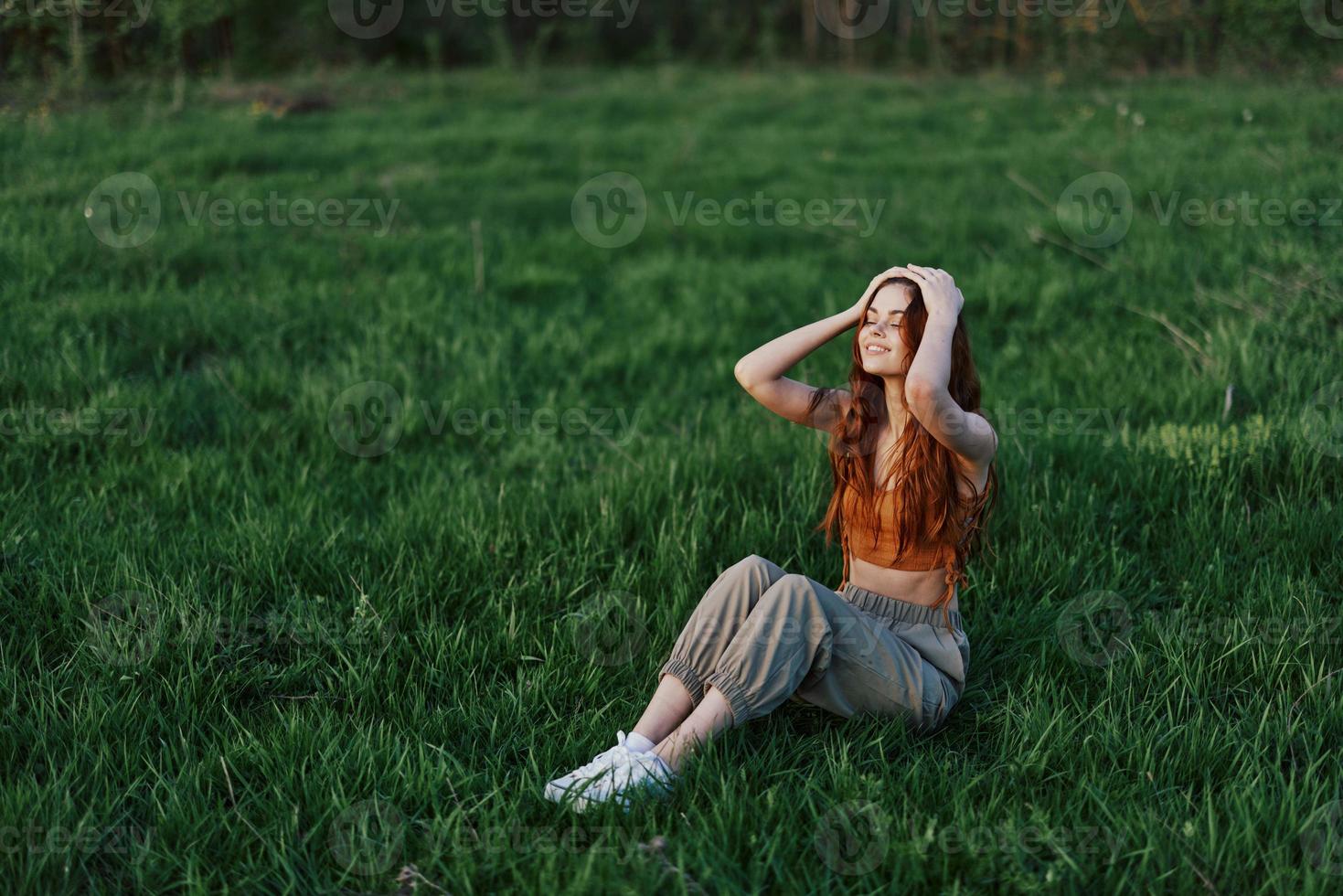 une roux femme avec long, venteux cheveux est assis en plein air sur le herbe dans le parc et sourires, le le coucher du soleil lumière éclairant sa affronter. le concept de harmonie avec la nature en plein air photo