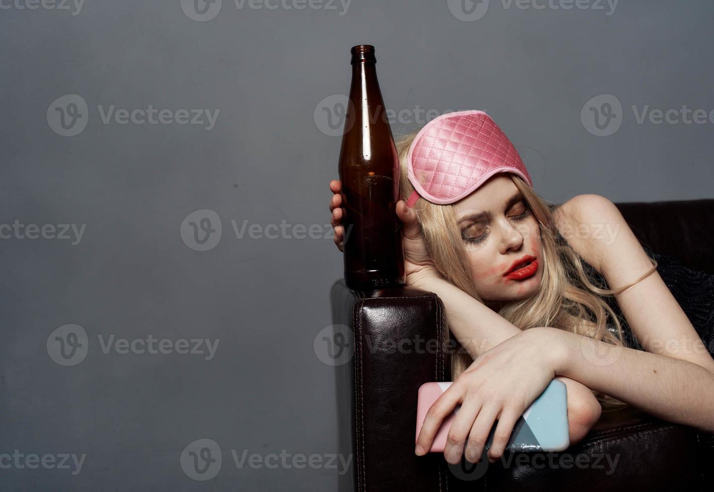 ivre blond femme avec une bouteille de Bière dort sur le canapé à l'intérieur sur une gris Contexte et une sommeil masque sur sa tête photo