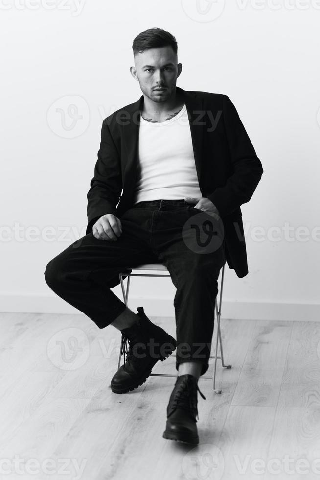 la modélisation instantanés. sérieux confiant attrayant Beau homme dans noir veste séance sur chaise posant dans blanc studio Contexte. noir et blanc concept. copie espace photo