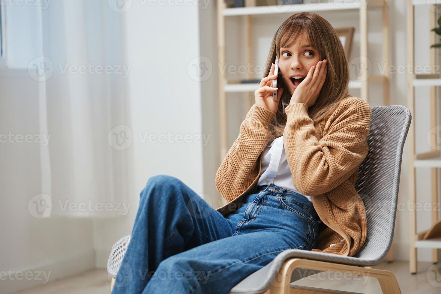 excité content surpris Jeune blond Dame dans chaud chandail avoir bien nouvelles de appel utilisation téléphone séance dans fauteuil à maison. pause de travail, prendre une casser, social médias dans gratuit temps concept. copie espace photo