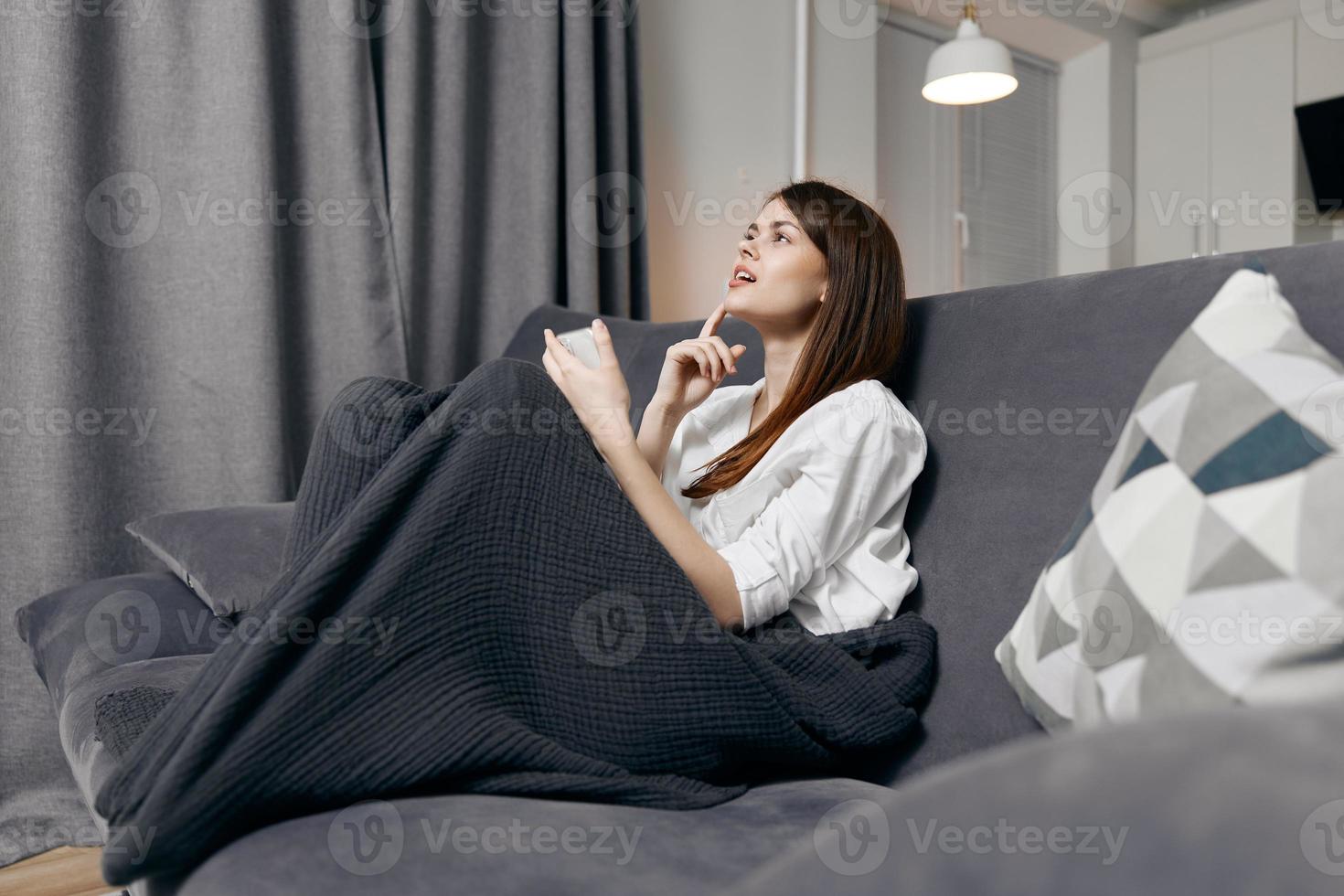 une femme est séance sur le canapé en dessous de une couverture et une mobile téléphone dans sa main photo