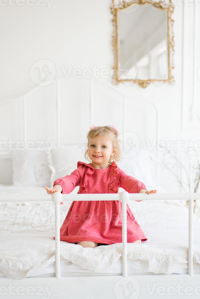 peu fille dans rose robe ayant amusement sur le lit dans le chambre photo