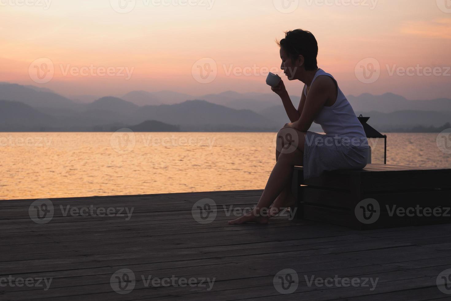 silhouette femme en portant tasse café séance par le Lac regarder lever du soleil. photo