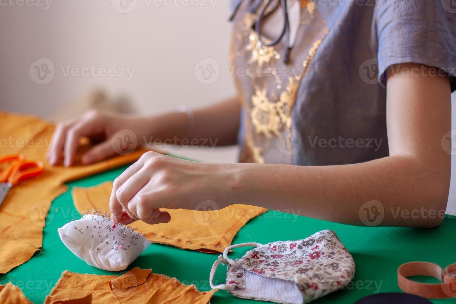 une Jeune fille couturière prend une aiguille de une oreiller pour aiguilles. Orange tissu sur une vert tableau. concentrer sur une tampon avec aiguilles et des doigts. proche mensonges une visage masque. horizontal. photo