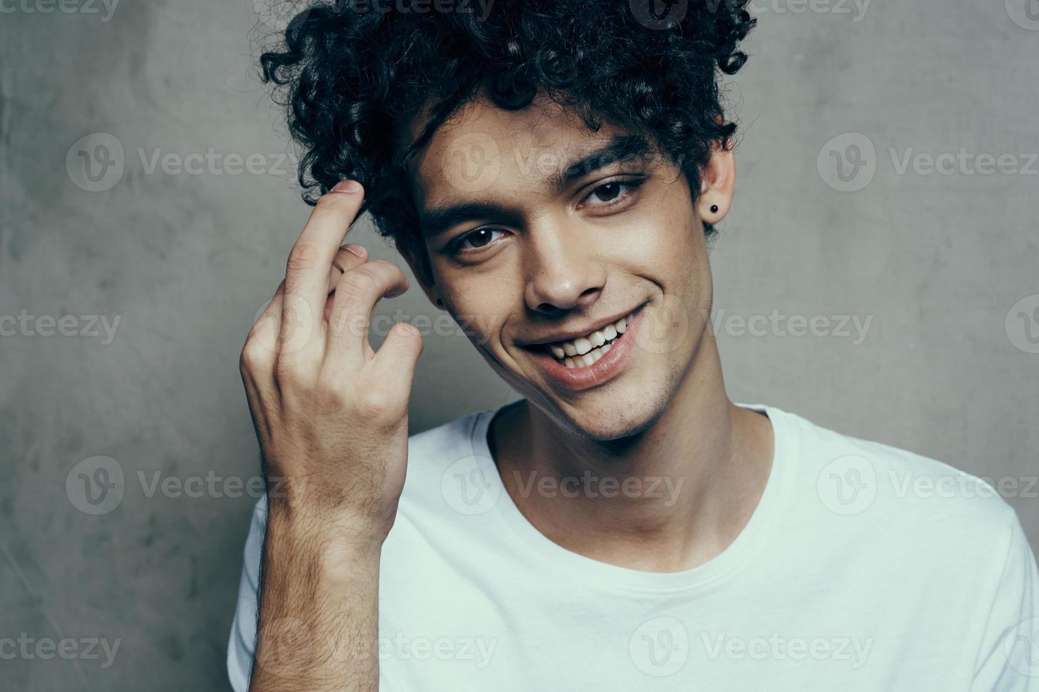 branché adolescent avec frisé cheveux dans une blanc T-shirt sur une gris Contexte amusement émotions photo