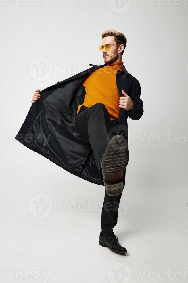 à la mode homme dans une manteau et pantalon est dansant sur une lumière Contexte et un Orange chandail des lunettes photo