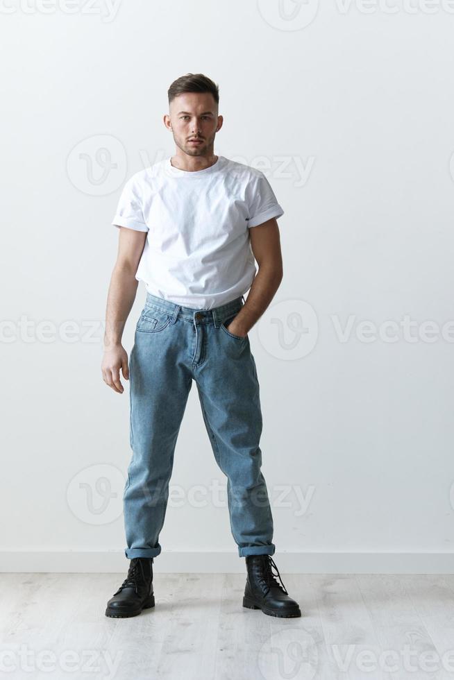 plein corps coup de Beau sérieux bronzé homme gars dans de base T-shirt détient main sur jeans posant sur blanc Contexte. mode style Nouveau collection offre. copie espace pour publicité. la modélisation instantanés photo